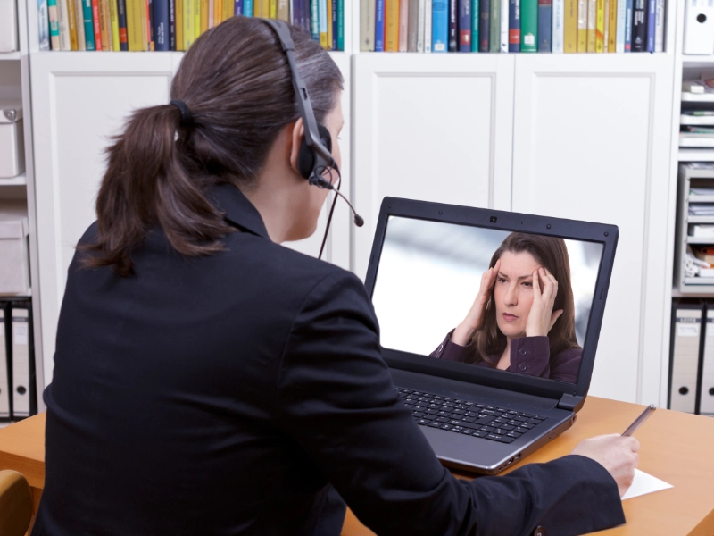 Photo angled looking over the shoulder of a therapist with headphones on and patient displayed on laptop screen.